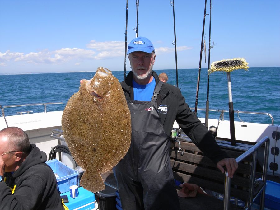 A man wearing fishing overalls and a blue cap holding a large flat fish aboard a boat at sea.