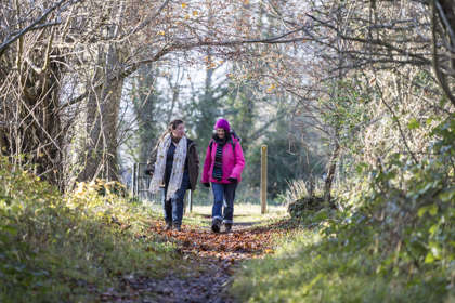 Two women walking along a woodland path, one wearing a pink coat and hat, both wearing walking boots.