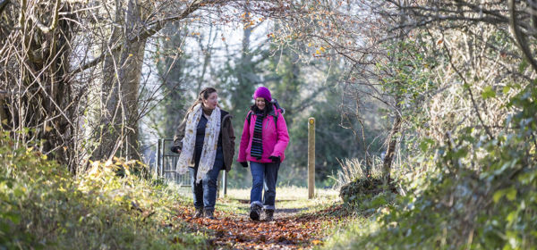 Two women walking towards the camera along a woodland path, one wearing a pink coat and hat, both wearing walking boots. The trees are bare and there are leaves on the path.