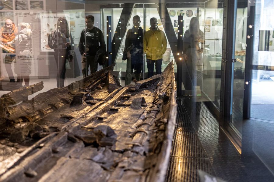 Part of a wooden boat in a glass case with people looking on