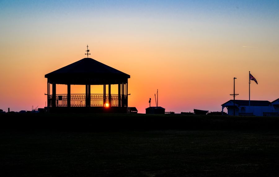 Silhouette of Deal Memorial Bandstand, boats and beach huts against a sunrise sky.