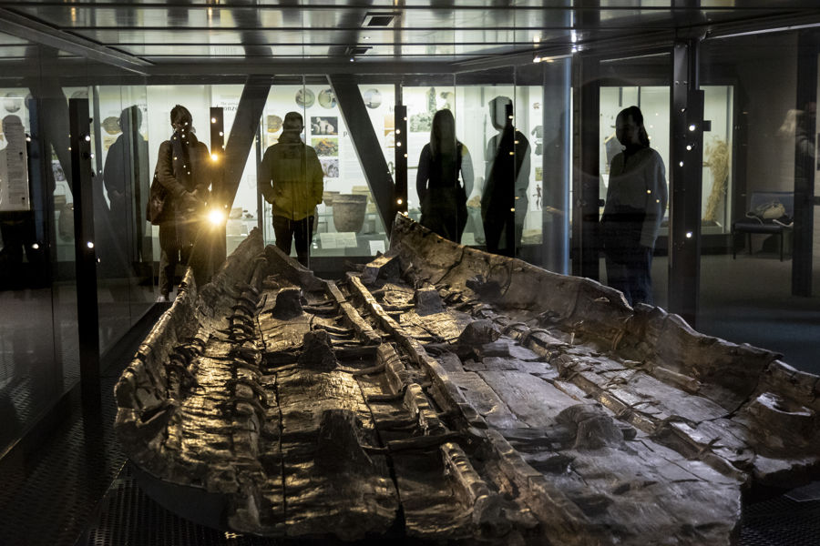 The remains of a large wooden boat in a large glass case with people looking in