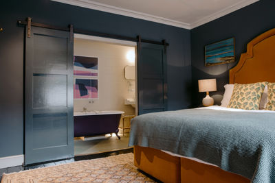 A bedroom with mustard coloured bed, dark grey walls and open door revealing a roll-top bath in the en-suite bathroom. 