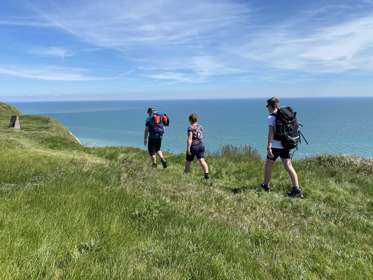 Three walkers carrying rucksacks walking along the cliff edge with blue sea and blue sky in the background.