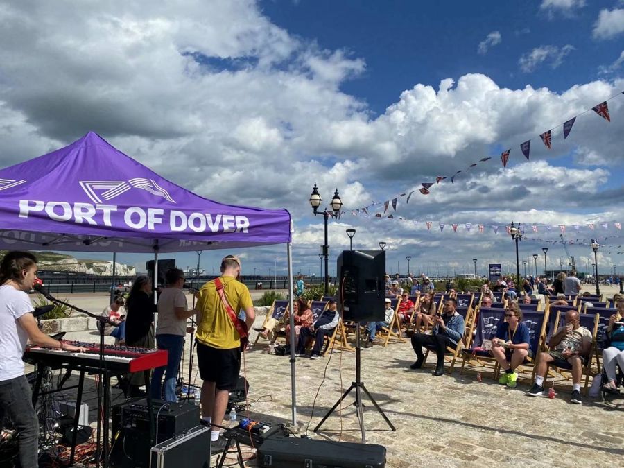 A crowd of people in deckchairs and a band playing under a gazebo on Dover Marina Curve.