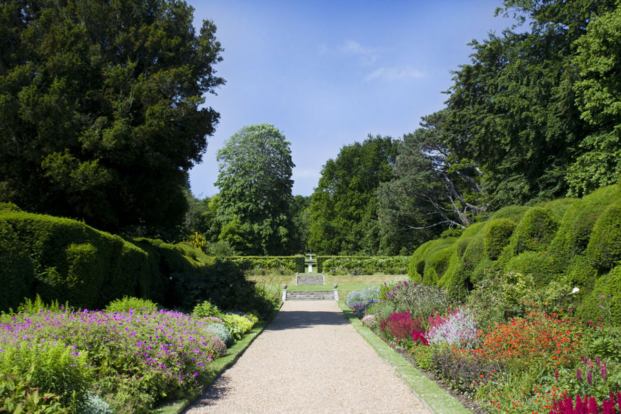 Gravel path bordered by flowerbeds, yew hedges and towering trees.