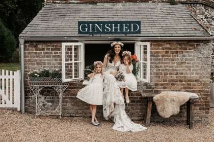 A smiling bride with two small bridesmaids sitting in a window of a brick gin shed.