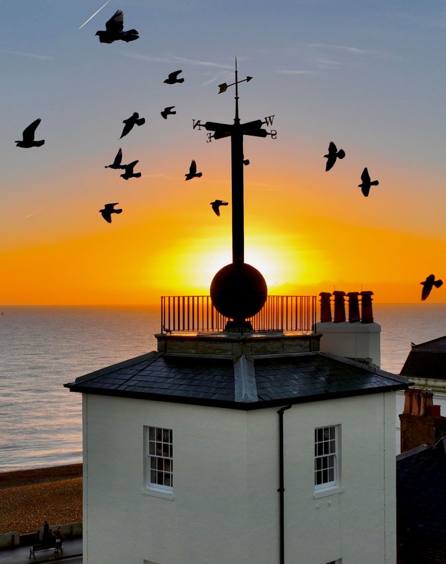 The sun rising behind the timeball tower and a flock of birds silhouetted against the sky.